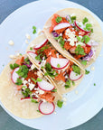 Salmon taco with radishes and fresh herbs and cheese on a white plate over a grey background.