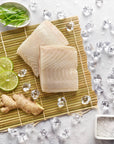 Wild sablefish fillets on a sushi mat with fresh ginger, lime slices, a bowl of salt and ice on a light marble background