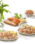 Assorted seafood dishes including salmon, scallops, and crab on a white surface with herbs and lemons.
