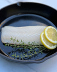 Raw halibut fillet in a cast iron pan with lemon slices and fresh thyme on a white background.