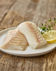 Two pieces of raw fish on a white plate with a lemon wedge and thyme sprigs on a wooden surface.