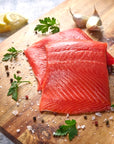 Two portions of sockeye salmon on a cutting board with fresh herbs, salt and pepper, garlic cloves and lemon slices in the background.